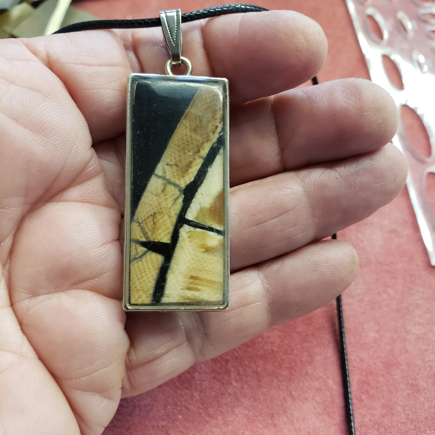 A hand holding a necklace with a rectangular pendant made of fossil mammoth ivory with visible black and beige patterns against a red background.